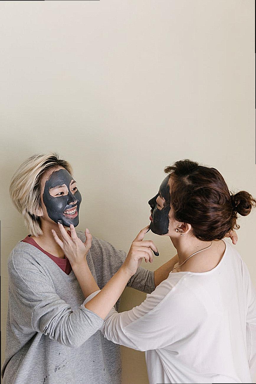 Playful girlfriends with facial mud mask touching chins of each other on beige background