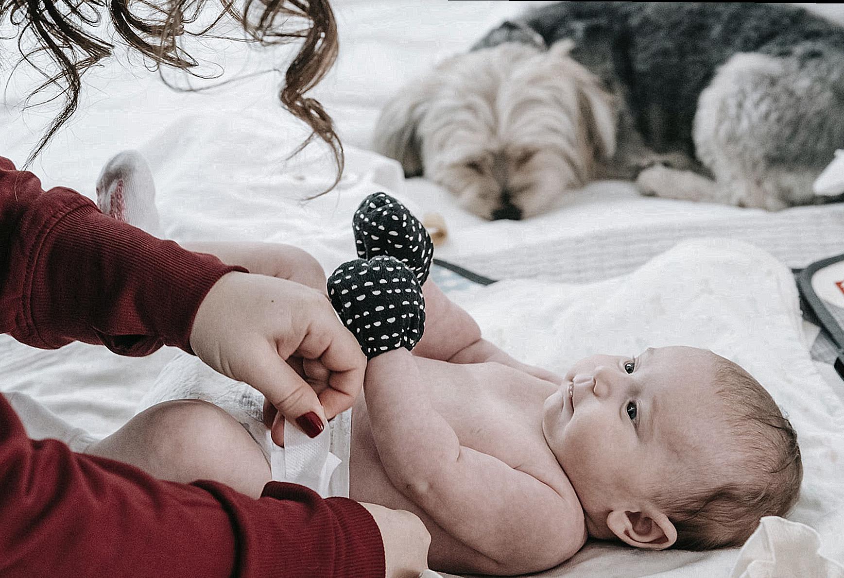 Crop anonymous mother putting on diaper on adorable infant baby wearing anti scratching mittens while lying on comfortable couch near hairy dog in light room