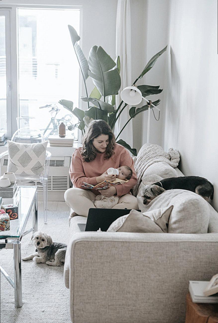 Caring mother feeding baby with bottle while sitting on sofa with laptop in modern room with dog and potted plant