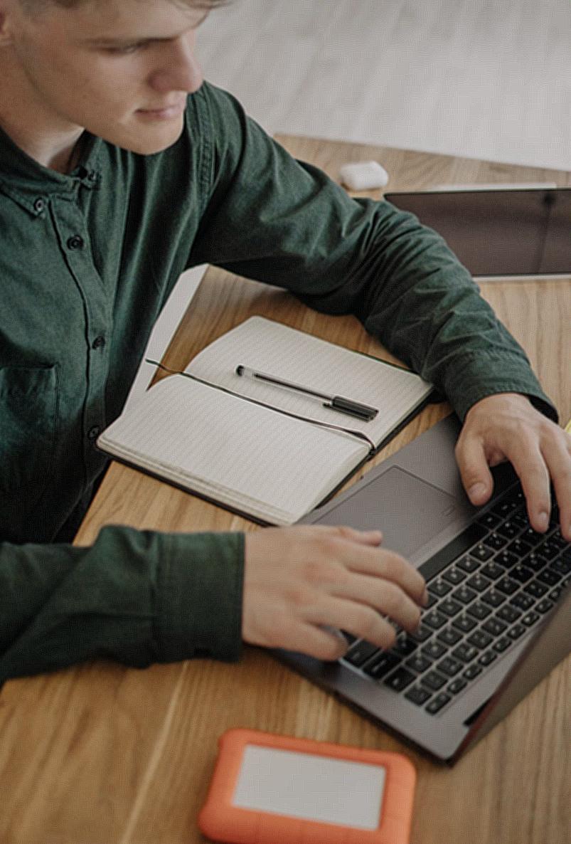 Close-Up Shot of a Male Student in Green Long Sleeves Using a Laptop on a Wooden Desk