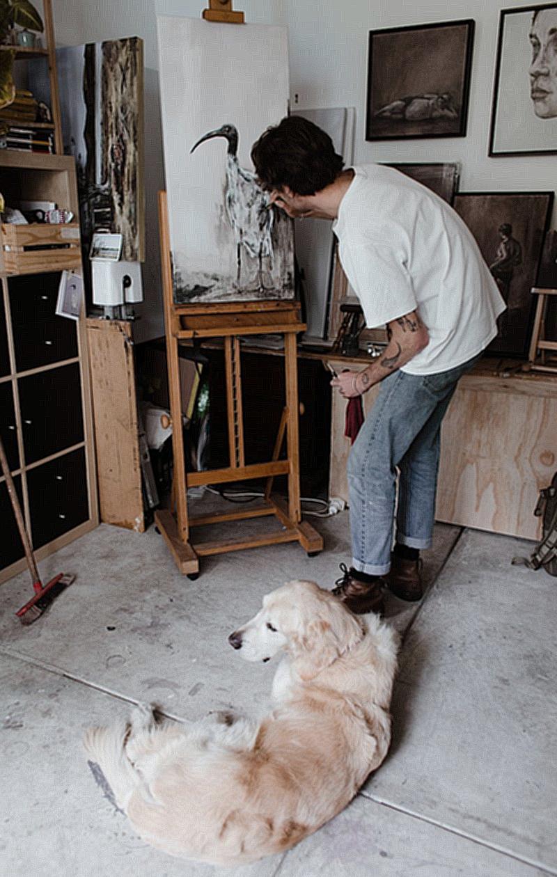 Side view of unrecognizable young male artist in casual clothes painting on easel while standing in light workshop near adorable Golden Retriever dog