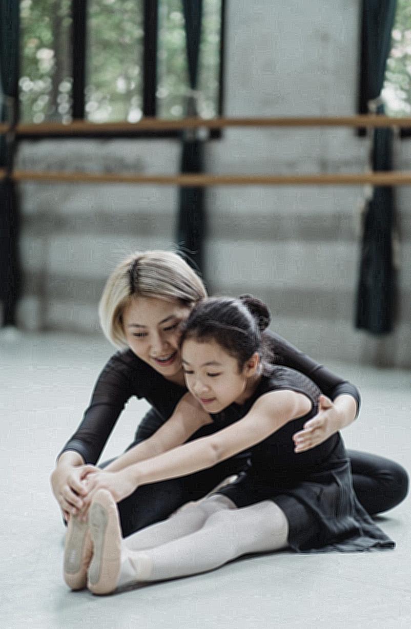 Full body positive Asian female ballet instructor sitting on floor and helping ballerina girl to stretch body in modern ballet school