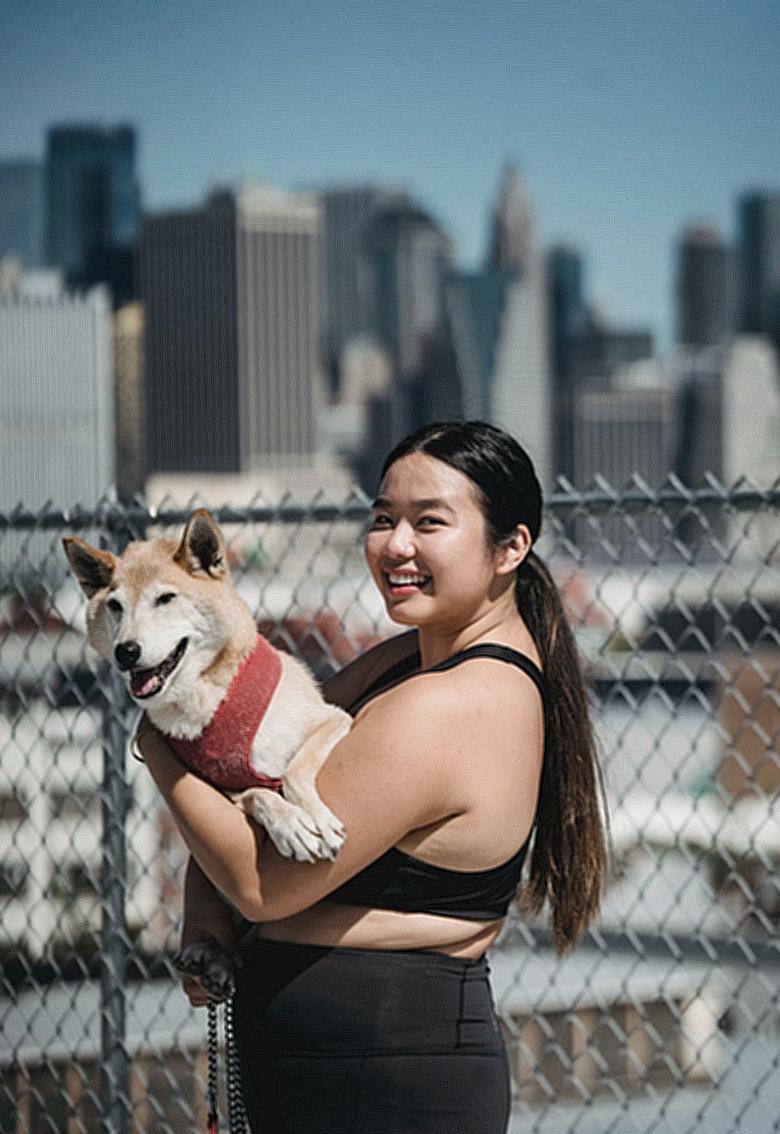 Side view of young ethnic female in sportswear standing near lattice fence and hugging cute puppy while looking at camera