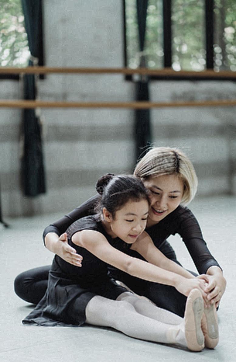 Full body positive Asian female ballet instructor sitting on floor and helping ballerina girl to stretch body in modern ballet school