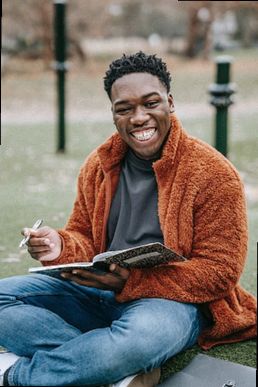 Smiling African American male student with copybook looking at camera while sitting on grassy lawn during homework preparation on blurred background